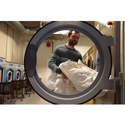 En mand i en stribet sweater lægger hvidt sengetøj i en vaskemaskine, set inde fra maskinen i et vaskeri.