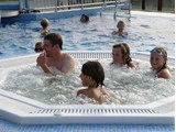 Flere børn og en voksen sidder i et boblende spabad, mens andre svømmer og leger i poolen omkring dem på en solrig dag.