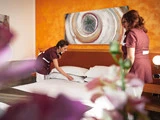 To stuepiger i uniformer reder en pænt opredt seng i et lyst indrettet hotelværelse med orange vægge og abstrakt kunst over hovedgærdet.