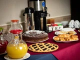 En morgenmadsbuffet med juicekander, en kaffeurne, en chokoladekage, en frugttærte, croissanter og teposer på et bord med en rød dug.