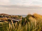 En kystby med huse med røde tage, grønne græsklædte bakker i forgrunden og havet synligt under en overskyet himmel i baggrunden.