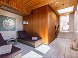 Moderne spa-rum med træsauna, to flettede liggestole med håndklæder, klinkegulv, ovenlys og kunstværker på væggen.