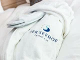 En hvid badekåbe broderet med DER SEEHOF og et blåt svanelogo, med et par tøfler pakket ind i plastik placeret ovenpå.