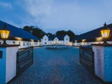 En hvid herregård med sorte tage, set gennem åbne smedejernsporte i skumringen, med varme lanternelys og en grusbelagt indkørsel, der fører til indgangen.