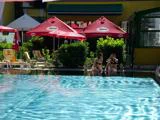 To kvinder i bikini sidder og taler ved en pool med røde parasoller og liggestole i baggrunden på en solrig dag.