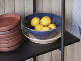 En blå skål fyldt med citroner står oven på en hvid skål ved siden af en stak røde tallerkner på en sort hylde mod en lys trævægsbaggrund.