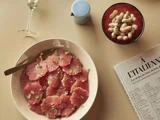 En tallerken med grapefrugt i tynde skiver med krydderurter, en skål med peanuts i skaller, et glas hvidvin, en lille blå kande og en del af en avis på et beige bord.