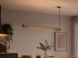 En moderne, rektangulær pendel hænger over et spisebord med en plante som bordpynt, skåle og blødt, varmt stemningslys i et minimalistisk rum.