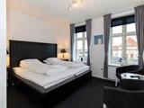 Et moderne hotelværelse med en dobbeltseng, to lamper, to vinduer med grå gardiner og to sorte stole omkring et glasbord.
