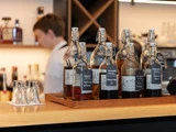 Flere mærkede glasflasker fyldt med forskellige væsker står på en træbakke ved en bardisk med en sløret bartender i baggrunden.