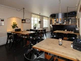 Et hyggeligt restaurantinteriør med træborde, sorte stole, stearinlys, planter og et barområde i baggrunden under moderne hængelamper.