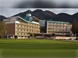 En moderne hotelbygning i flere etager med balkoner, der ligger på en baggrund af skovklædte bjerge og en græsmark i forgrunden.