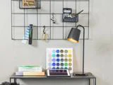 Et skrivebord med bøger, en åben tablet, en lampe og en farvepalet; bagved er der en metalgittervæg med kontorartikler og en lommeregner.