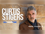 En mand med gråt hår og skæg, iført brun jakke og sort tørklæde, står op ad en stenmur. Teksten lyder: Curtis Stigers, All Things Live, Nørre Vosborg. Foto: Andy Lawless.