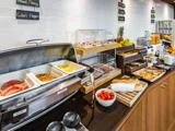 En morgenmadsbuffet med varme retter, pålæg, kager, brød, frugt, cherrytomater, juice og skilte med forskellige madvarer.