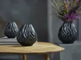 En sort, skulpturel vase med et bølget flammelignende design står på et træbord; en anden vase med blomster er sløret i baggrunden.