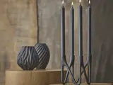 Fire tændte sorte bloklys i en moderne, skulpturel sort holder står på en træplade med to sorte vaser med struktur i baggrunden mod en neutral væg.