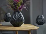 En sort, bølgemønstret vase med lilla og grønne blomster står på et træbord. To lignende vaser er slørede i baggrunden.