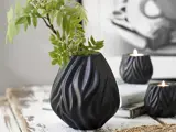 En sort, skulpturel vase med grønne bladgrene står på et rustikt hvidt bord ved siden af to matchende sorte lysestager med tændte stearinlys.