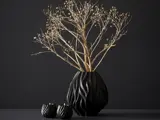 En sort vase med et bølget mønster indeholder tørre, bladløse grene. To små, matchende sorte lysestager med tændte stearinlys står ved siden af, alt sammen på en mørk baggrund.