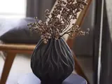 En sort, struktureret vase med dekorative, tørrede grene står på et mørkt, rundt bord i et svagt oplyst rum med en træstol i baggrunden.