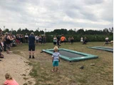 Folk samles udendørs omkring to trampoliner i jordhøjde, mens en ung dreng i forgrunden ser på. Det er overskyet, og omgivelserne ser ud til at være en park eller et åbent græsareal.
