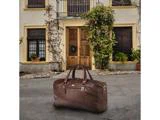 En stor brun lædertaske ligger på fortovet foran et hus med trædøre, potteplanter og klatrende slyngplanter.