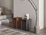 Minimalistisk interiør med betontrapper, to kantede skamler (brune og sorte), en abstrakt skulptur, en vase med en bar gren og et geometrisk tæppe på et poleret gulv.