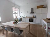 Et lyst køkken med hvide skabe, et træbord med fire stole, en blomsterdekoration i midten, hængende lamper og et stort vindue.