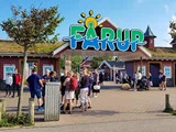 Folk samles ved indgangen til Fårup forlystelsespark under et farverigt skilt med træer og bygninger i baggrunden på en solrig dag.