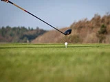 Et nærbillede af en golfkølle, der forbereder sig på at ramme en golfbold på en tee, med en græsklædt bane og træer i baggrunden.