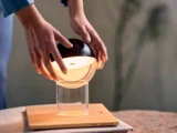En person placerer en moderne, rund lampe med sort top og glasbund på et gennemsigtigt, cylindrisk stativ, der står på en træfod.