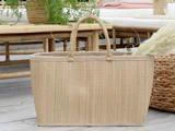 En vævet beige tote bag med håndtag ligger på et trægulv nær kurvemøbler og grønne planter i et hyggeligt, lyst rum.