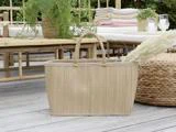 En flettet kurv med håndtag står på en træterrasse omgivet af havemøbler, grønne planter og sammenfoldede tæpper.