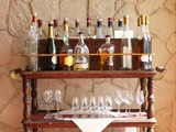 En trækærre, der står op ad en stenvæg, har mange flasker spiritus på den øverste hylde og flere tomme vin- og cocktailglas på den nederste hylde.