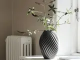 En mørkegrå, spiralformet vase med grene med blade står på en hvid vindueskarm ved siden af en radiator.