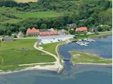 Luftfoto af en lille lystbådehavn med både, en parkeringsplads, bygninger med røde tage og grønne marker ved vandkanten.