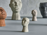 Fem skulpturelle hovedbuster i forskellige størrelser og farver er arrangeret på en grå overflade mod en struktureret grå baggrund.