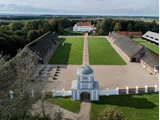 Luftfoto af et stort gods med en hvid herregård, grønne plæner, murstensbygninger og en central grussti, der fører til en hvid port under en kuppelformet struktur.