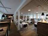 Moderne restaurantinteriør med trægulve, hvide vægge, borde dækket af hvide duge, sorte stole, hængelamper og naturligt lys fra store vinduer.