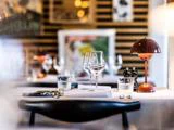 Nærbillede af et restaurantbord dækket med vinglas, vandglas, tallerkener, bestik, en rød lampe og salt- og peberbøsser med abstrakt kunst på væggen i baggrunden.