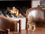 To glas champagne på et træbord, med brune læderstole og en tændt pejs i baggrunden.