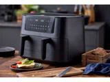 En sort frituregryde med to kurve og et digitalt display står på et køkkenbord af træ ved siden af en tallerken med mad, brød og køkkenredskaber.