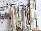 Flere stribede og ensfarvede håndklæder hænger på kroge på en sort stang op ad en hvid murstensvæg med en træstige og flettede kurve i nærheden.