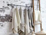 Flere stribede og ensfarvede håndklæder hænger på kroge på en sort stang op ad en hvid murstensvæg med en træstige og flettede kurve i nærheden.
