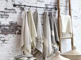 En række stribede og ensfarvede neutralfarvede håndklæder hænger på kroge på en sort stang mod en hvid murstensvæg med en træstige og flettede kurve i nærheden.