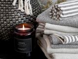 Et tændt linnedduftlys står ved siden af en stak mønstrede, foldede håndklæder og en vævet kurv med frynser i et hyggeligt miljø.