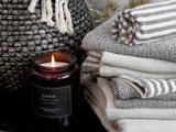 Et tændt linnedduftlys står ved siden af en stak mønstrede, foldede håndklæder og en vævet kurv med frynser i et hyggeligt miljø.