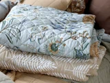 To foldede quilts stablet oven på hinanden; den øverste quilt har et blomstermønster, og den nederste quilt har en beige farve med hvide bladmotiver.