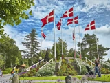 Flere danske flag på høje stænger står blandt blomsterbede og grønt i en park, og folk går rundt i nærheden under en delvist overskyet himmel.